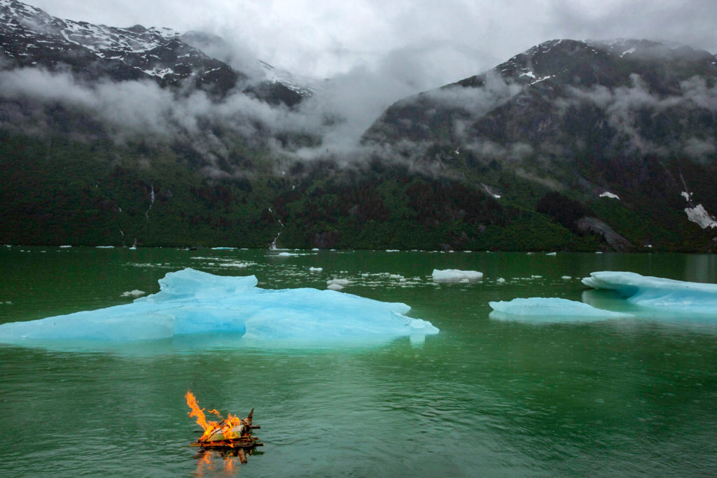 Cremains on a driftwood raft sail across jade waters towards a turquoise ice berg. Clouds shroud the mountains in the distance. Image copyright amandacastleman.com