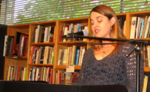 Amanda Castleman reading at Seattle's University Book Store (a pale white lady speaking into a mic, standing in front of a packed bookcase. She has long brown hair parted on the side, and is wearing a loose-knit sage v-neck sweater.)