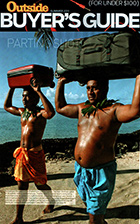 A shirtless Samoan man and boy carry luggage on their heads. They're on a sandy beach with dark lava rocks along the shoreline, and turquoise sea and sky behind