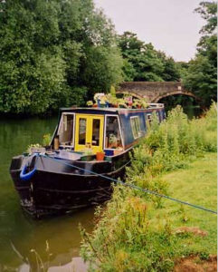 A pastoral British canal scene. A navy narrowboat is moored in front of an arched brick bridge. It has bright yellow doors and colorful plants on its roof.