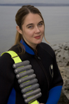 An image of Amanda Castleman about to cold-water dive. At the time of the image, she was in her mid-30s. A pale white woman with dark hair pulled into a low pony. She's wearing a wetsuit and has a homemade dive belt over her shoulder, which sort of looks like a bandolier made out of steel hot dogs. The day is overcast and she's standing on a rocky Pacific Northwest Beach with grey water visible in the background.
